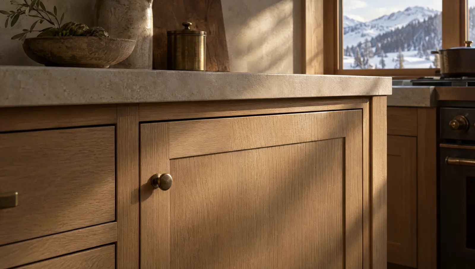 Close view of a white oak cabinet door with a round brass knob below a limestone counter, lit by low winter sun with snowy foothills in the window.